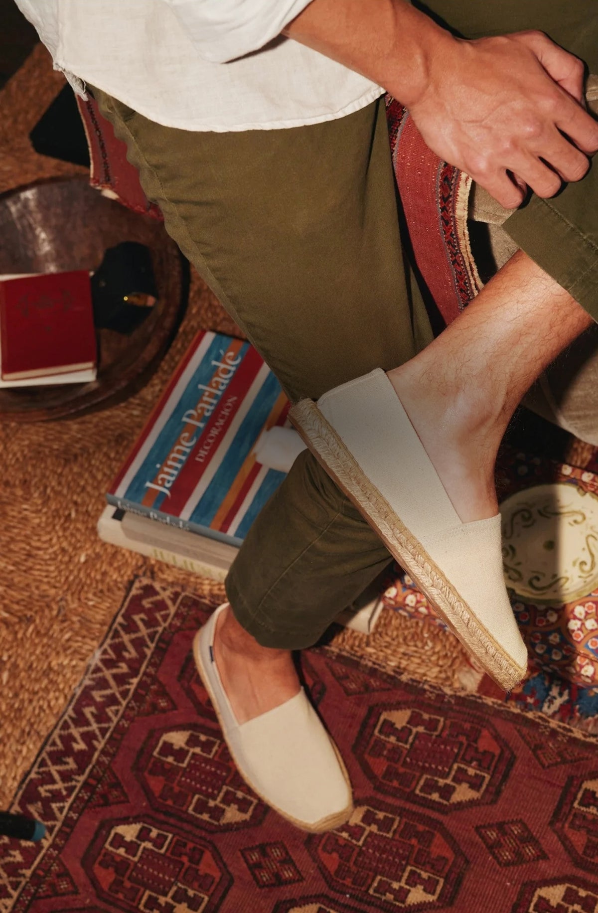 Soludos lifestyle image featuring a man wearing natural undyed canvas espadrilles with hand-stitched jute soles, styled with olive trousers in a relaxed indoor setting.